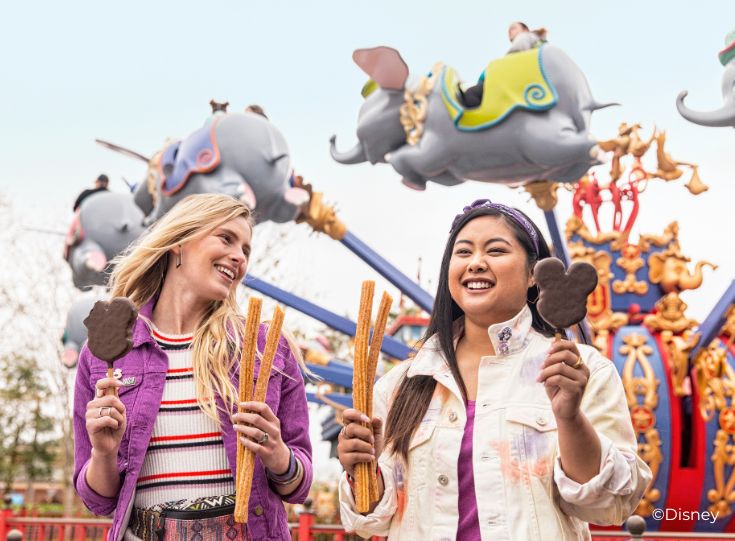 Two young ladies eating Mickey bars and churros in front of Dumbo ride. Two young ladies eating Mickey bars and churros in front of Dumbo ride.